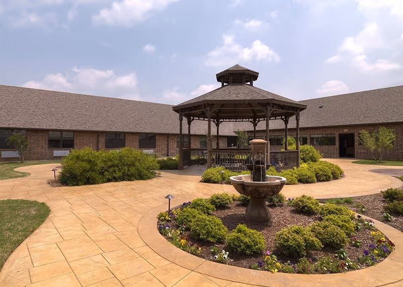 Courtyard with a central gazebo and fountain, paved walkways and landscaped shrubs in front of a single-story brick building under a partly cloudy sky.