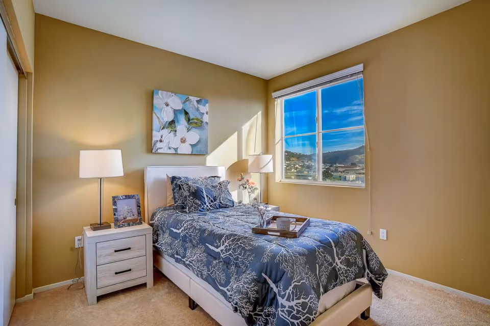 Sunlit bedroom with a single bed made with patterned bedding, a nightstand and lamp, floral artwork above the headboard, and a window showing hills outside.