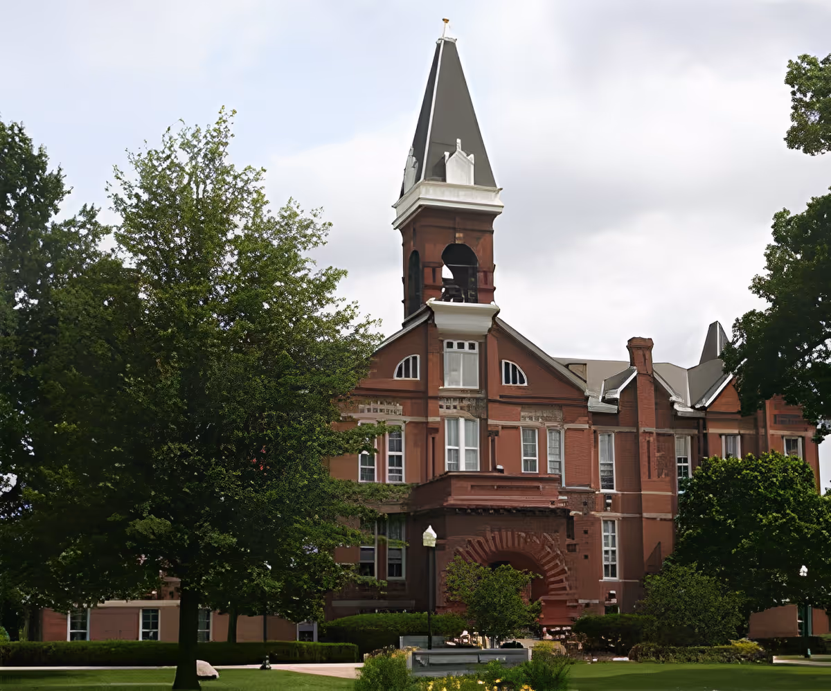 A large historic brick building with a tall pointed tower and arched entrance, surrounded by green trees and a well-maintained lawn under a cloudy sky.