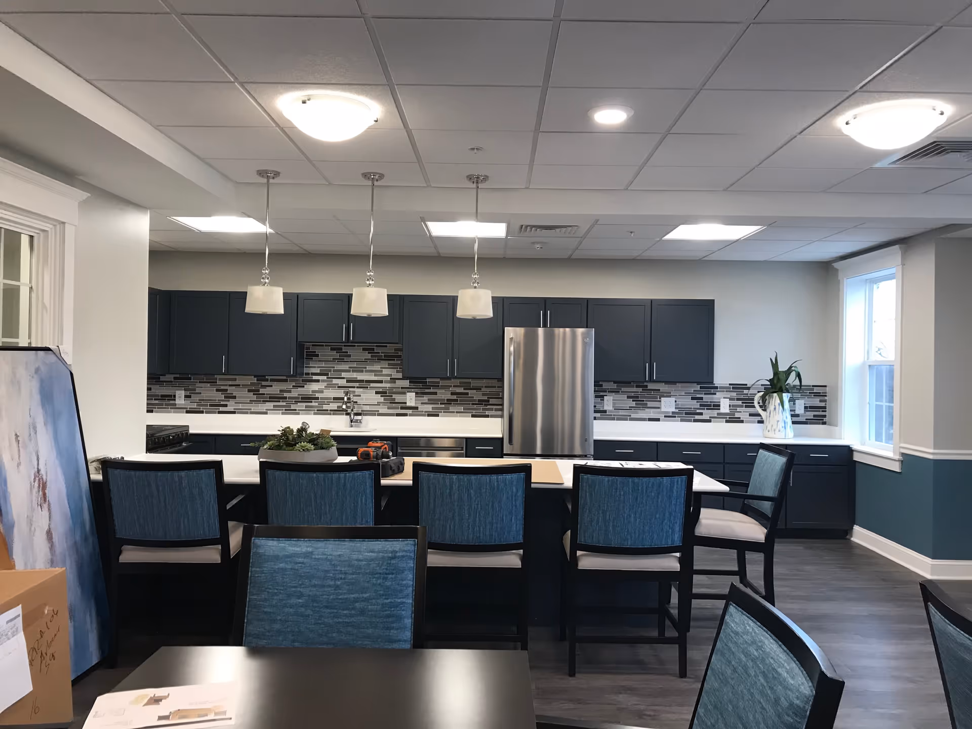 Modern kitchen area with dark blue cabinets, a stainless steel refrigerator, and a white countertop island with six blue cushioned chairs. The backsplash features a mosaic tile design in shades of gray and white. Three pendant lights hang above the island, and there is a window on the right side letting in natural light. The floor is a dark wood laminate, and there are some boxes and a painting leaning against the wall on the left side.
