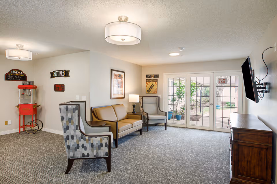 A cozy living room area in a memory and dementia care facility featuring a popcorn machine on the left, two patterned armchairs, a brown leather loveseat, a side table with a lamp, a wall-mounted TV on the right, and glass double doors leading to an outdoor garden area. The walls are decorated with vintage-style cinema signs.