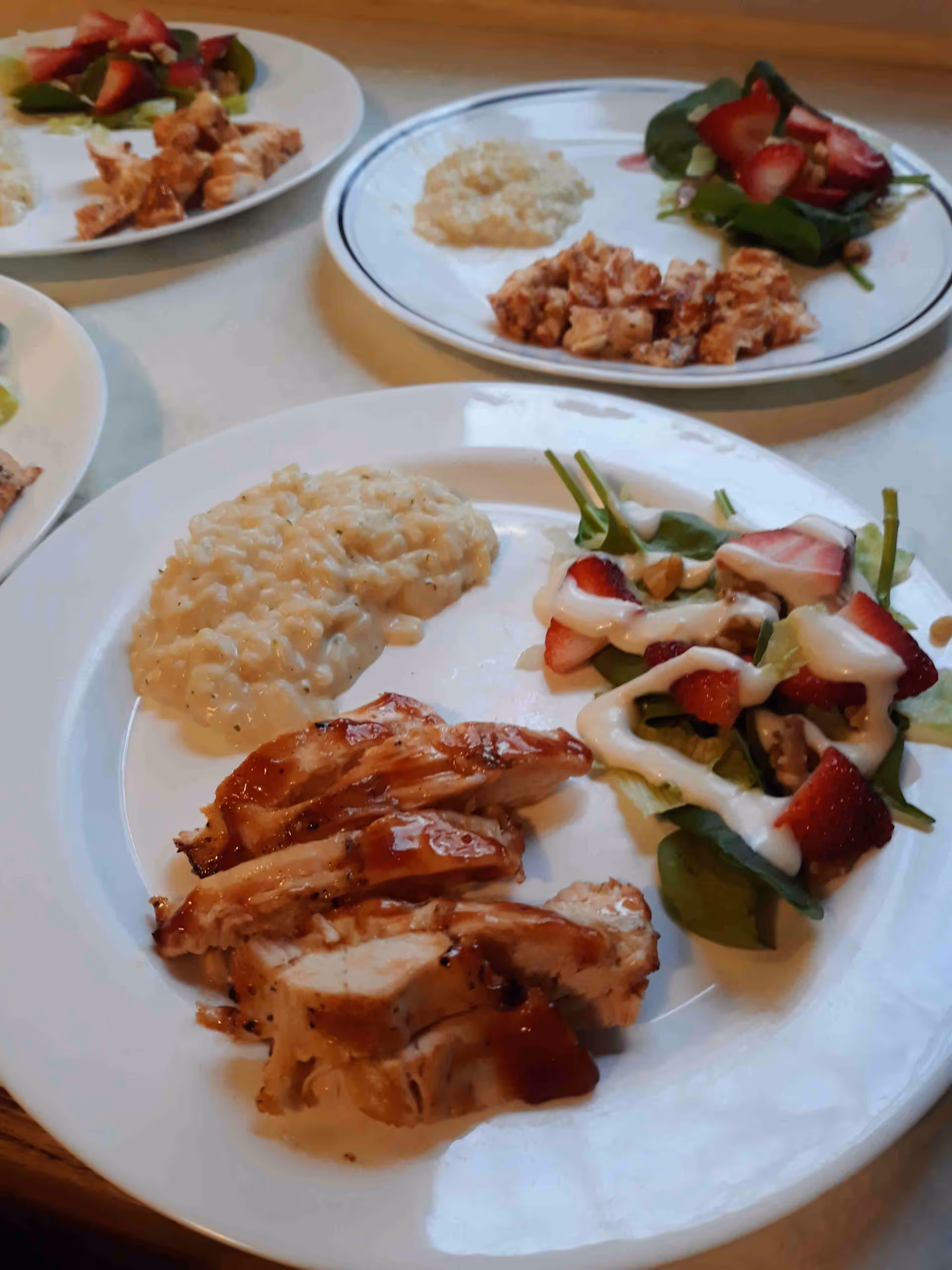 Plates of food on a table featuring grilled chicken, creamy risotto, and a fresh salad with spinach, strawberries, and dressing.