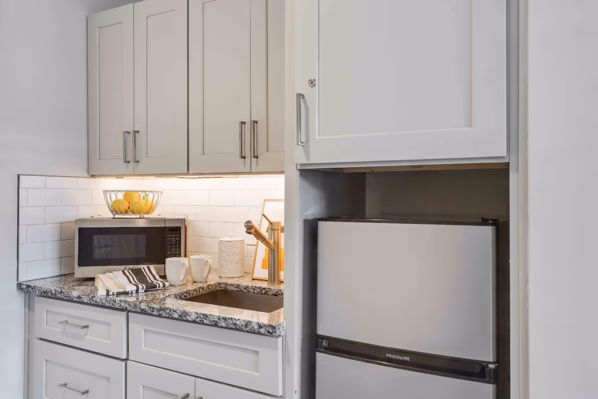 A small kitchen area featuring white cabinets, a granite countertop with a built-in sink, a microwave, two white mugs, a striped kitchen towel, a bowl of lemons, and a small refrigerator.