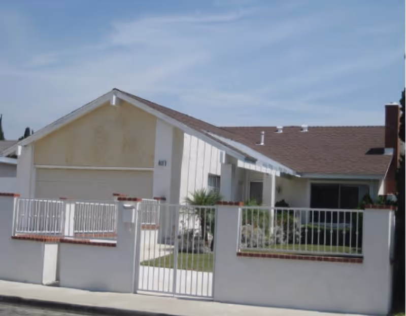 Single-story ranch-style house with an attached garage, gated front yard, and a low white fence under a blue sky.