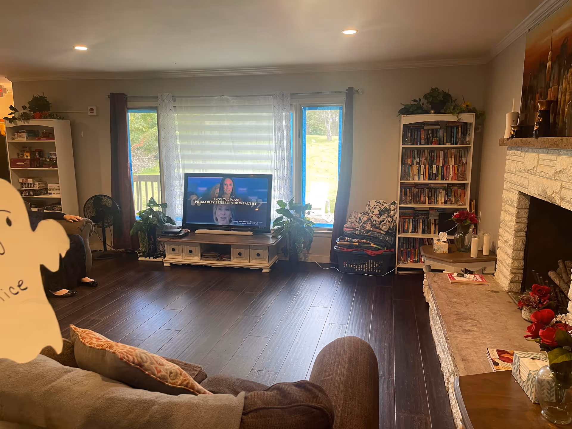 A cozy living room with dark wooden floors, a TV on a white stand showing a news program, two large windows with sheer and dark curtains, a bookshelf filled with books, a stone fireplace with candles and flowers on the mantel, and a brown couch with patterned pillows.