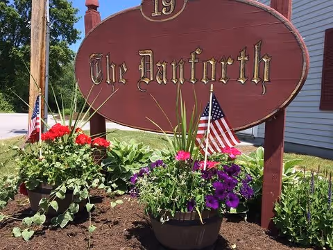 A large wooden sign reading 'The Danforth' with the number 19 above it, surrounded by flower pots containing colorful flowers and small American flags, situated outdoors next to a building and a street.