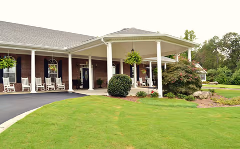 Exterior view of a senior living facility with a covered porch featuring white rocking chairs and hanging plants. The building has a brick facade and is surrounded by a well-maintained lawn and landscaped bushes.