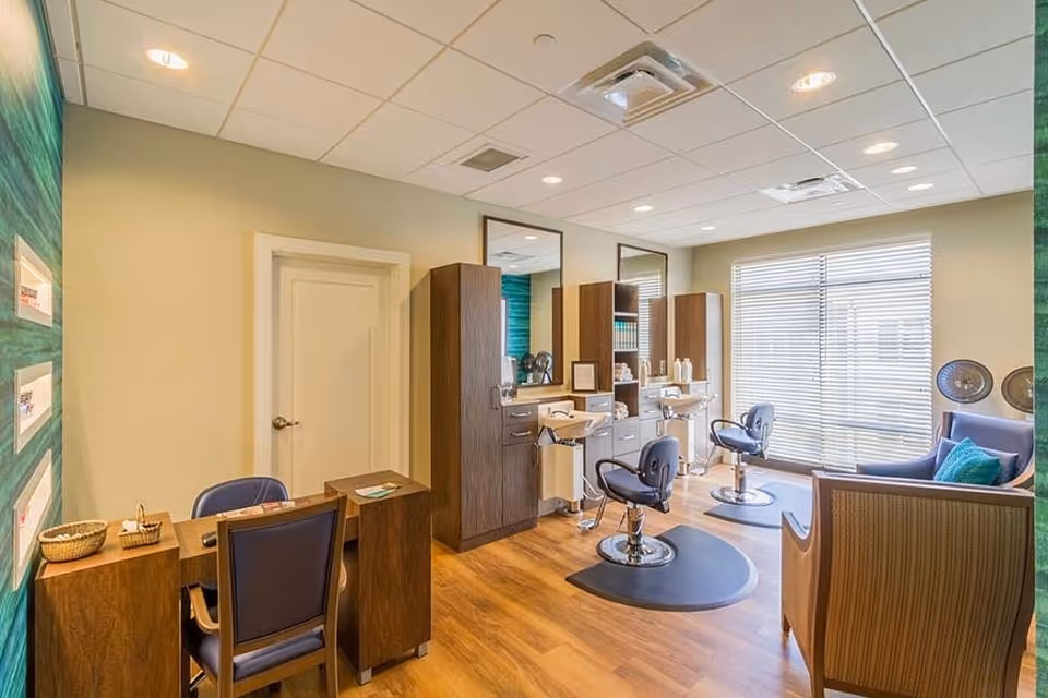 Bright salon-style room with styling chairs, shampoo sinks, mirrors, a small reception desk and seating by a large window.