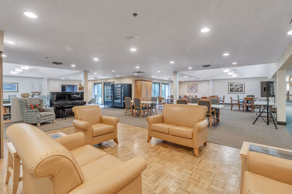 A spacious senior living facility common area with beige leather armchairs and a loveseat arranged on a parquet floor. In the background, there are multiple tables and chairs on a carpeted floor, a black upright piano, vending machines, and framed artwork on the walls. The ceiling has recessed lighting and the room has large windows letting in natural light.
