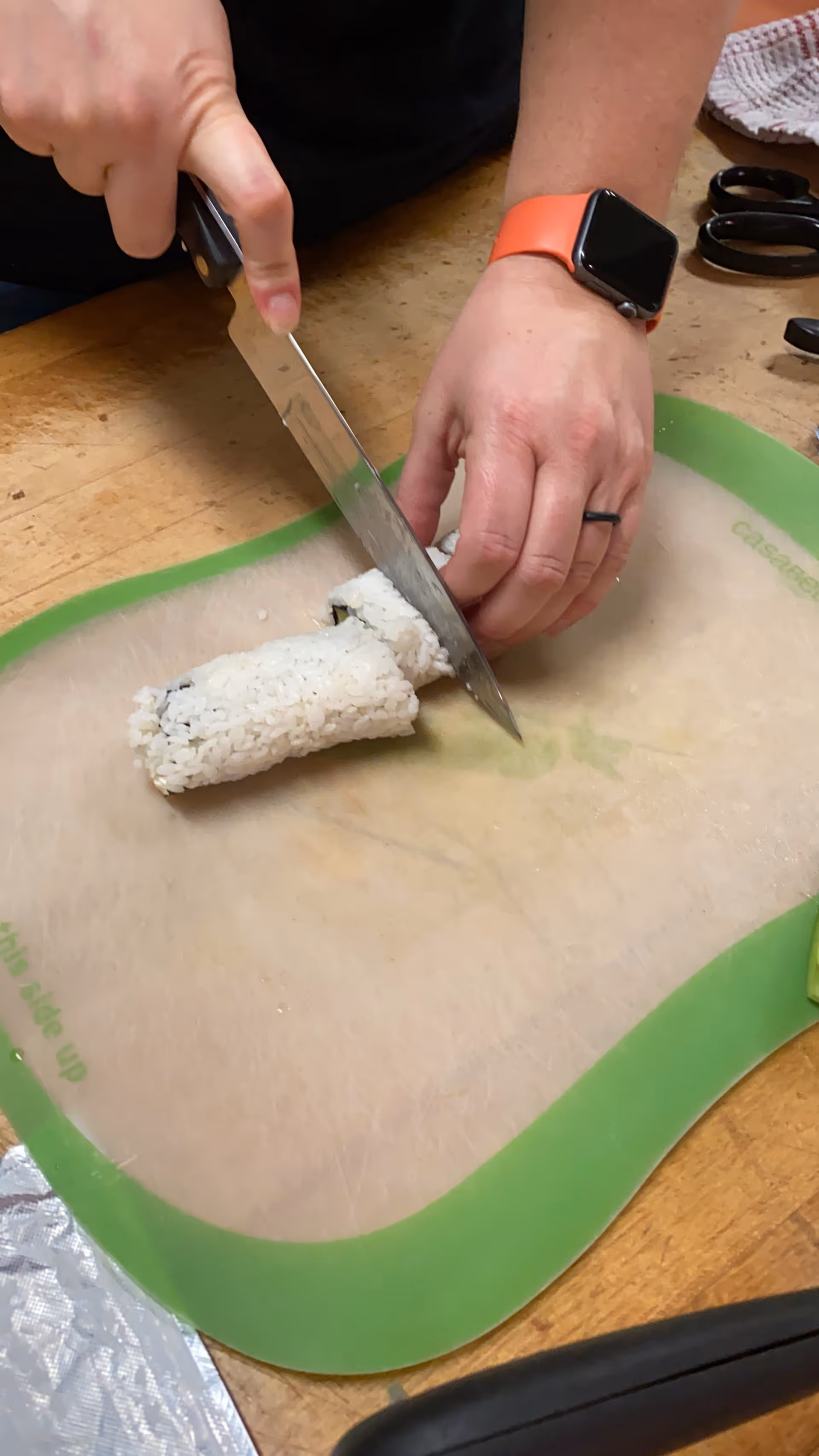 Hands cutting a sushi roll with a large knife on a green-edged cutting board.