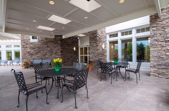 Outdoor covered patio area with metal tables and chairs, each table decorated with a flower pot. The space features stone walls, large windows, and ceiling lights.