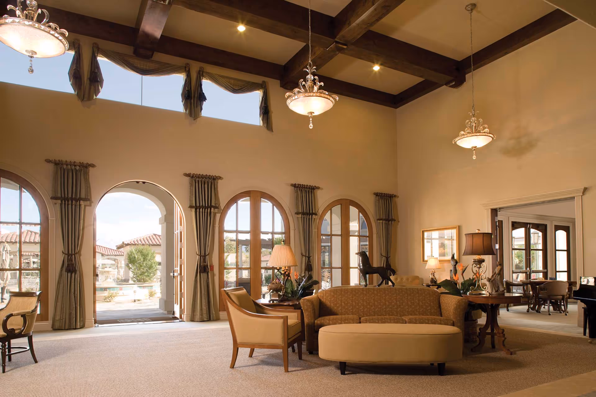 A spacious and elegant living room area with high ceilings featuring wooden beams and two ornate chandeliers. The room has large arched windows and doors with curtains, allowing natural light to enter and providing a view of an outdoor patio area. The seating includes a patterned sofa, an upholstered chair, and an ottoman, with side tables holding lamps and decorative items including a horse statue. In the background, there is another room visible with more seating and a piano.