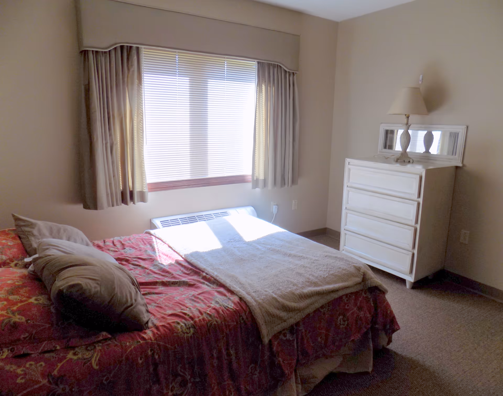 A bedroom with a bed covered in a red patterned bedspread and several pillows. There is a window with closed blinds and beige curtains. A white dresser with a lamp and a mirror on top is positioned against the wall. The room has beige walls and carpeted floor.