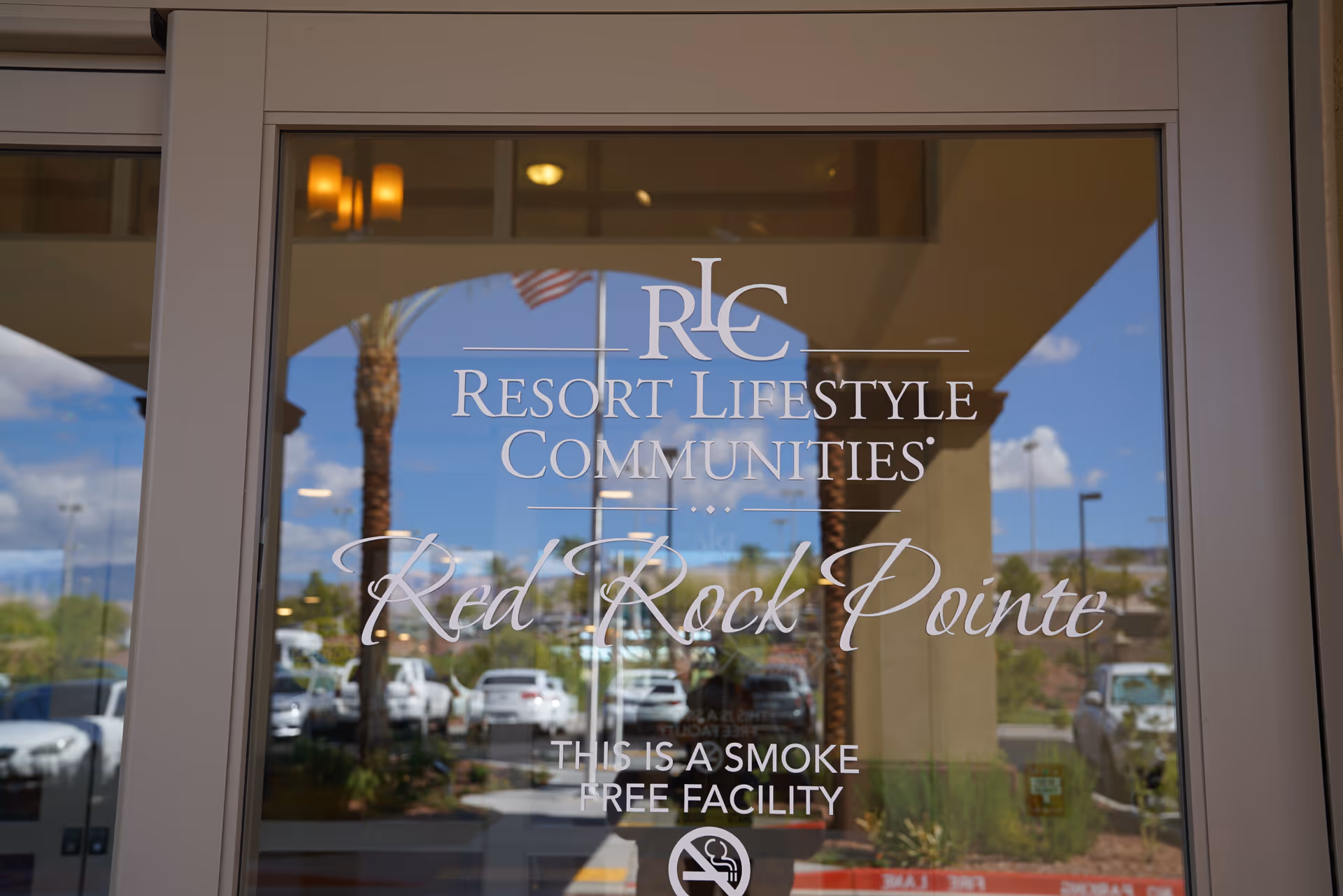 Glass door entrance with the text 'RLC Resort Lifestyle Communities Red Rock Pointe This is a smoke free facility' printed on it, reflecting a parking lot with cars, palm trees, and a blue sky with clouds.