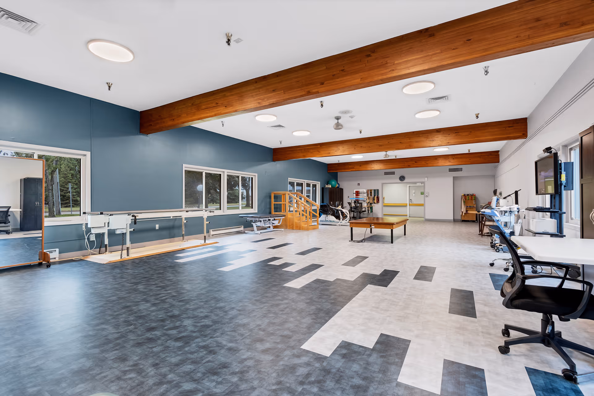 A spacious rehabilitation or physical therapy room with large windows, wooden ceiling beams, and a patterned floor. The room contains parallel bars for walking exercises, a large mirror, exercise equipment, a table, and chairs. The walls are painted blue and white, and the room is well-lit with ceiling lights.