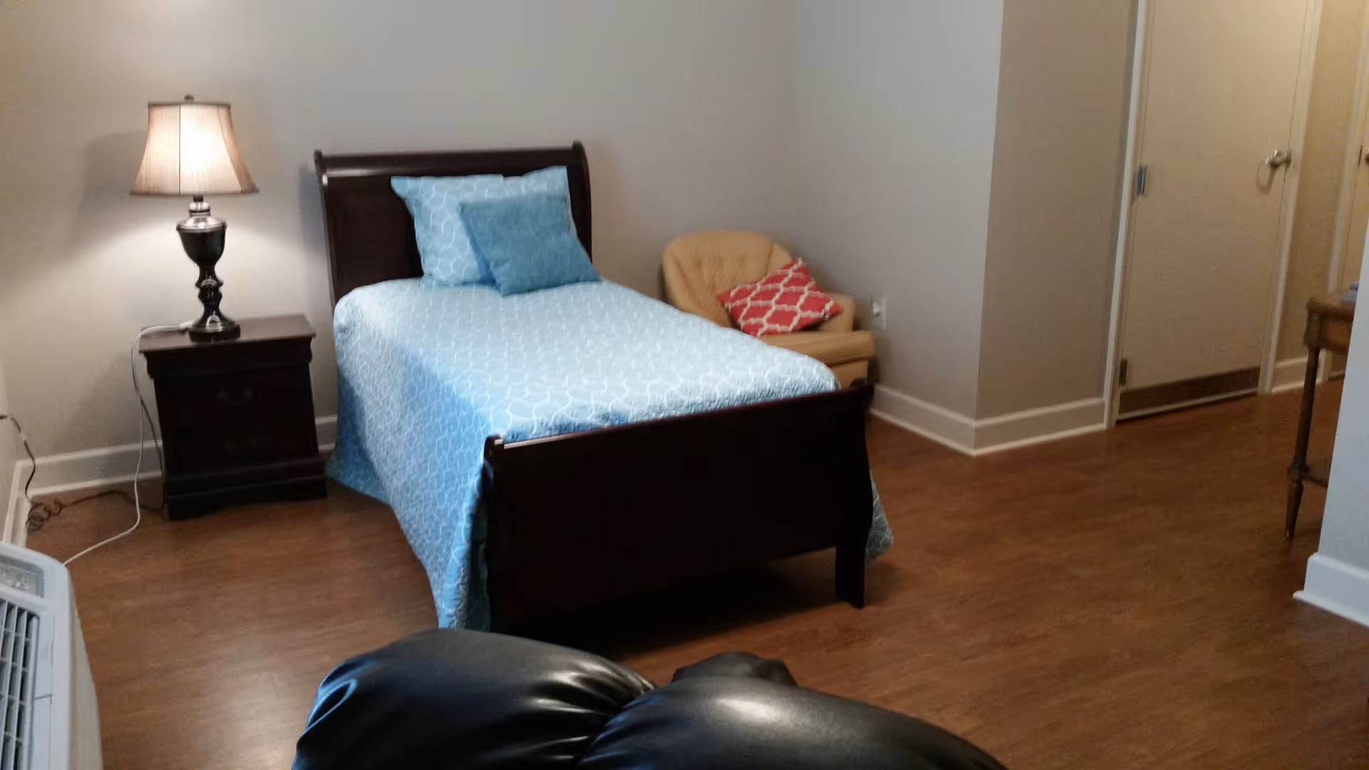 A bedroom with a single bed covered in a light blue patterned bedspread and two pillows. Next to the bed is a dark wooden nightstand with a lamp on it. There is a beige armchair with a red patterned cushion in the corner. The floor is wooden, and there is a door visible in the background.