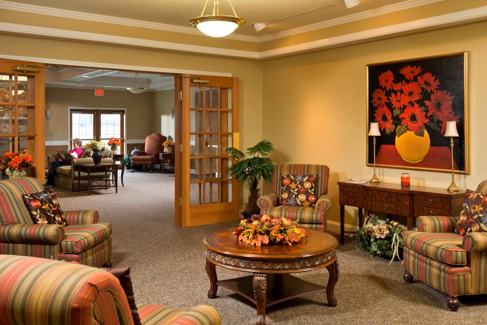 A cozy living room area in a senior living facility with striped armchairs featuring floral cushions, a round wooden coffee table decorated with a fall-themed centerpiece, and a wooden sideboard with two lamps and a large painting of red flowers in a yellow vase on the wall. In the background, through open wooden French doors, another seating area is visible with more armchairs and a person reading.