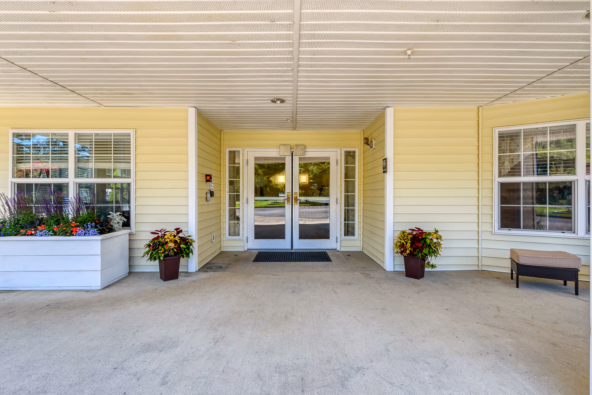 Covered entrance area of a building with double glass doors in the center, flanked by windows on both sides. There are flower pots with colorful plants on either side of the doors and a bench with a cushion on the right side. The exterior walls are light yellow with white trim.