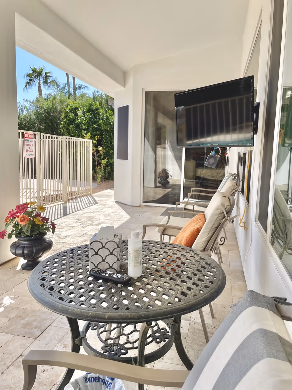 Covered outdoor patio at a senior living facility with a round metal table, cushioned chairs, a wall-mounted TV, potted flowers, and glass doors.