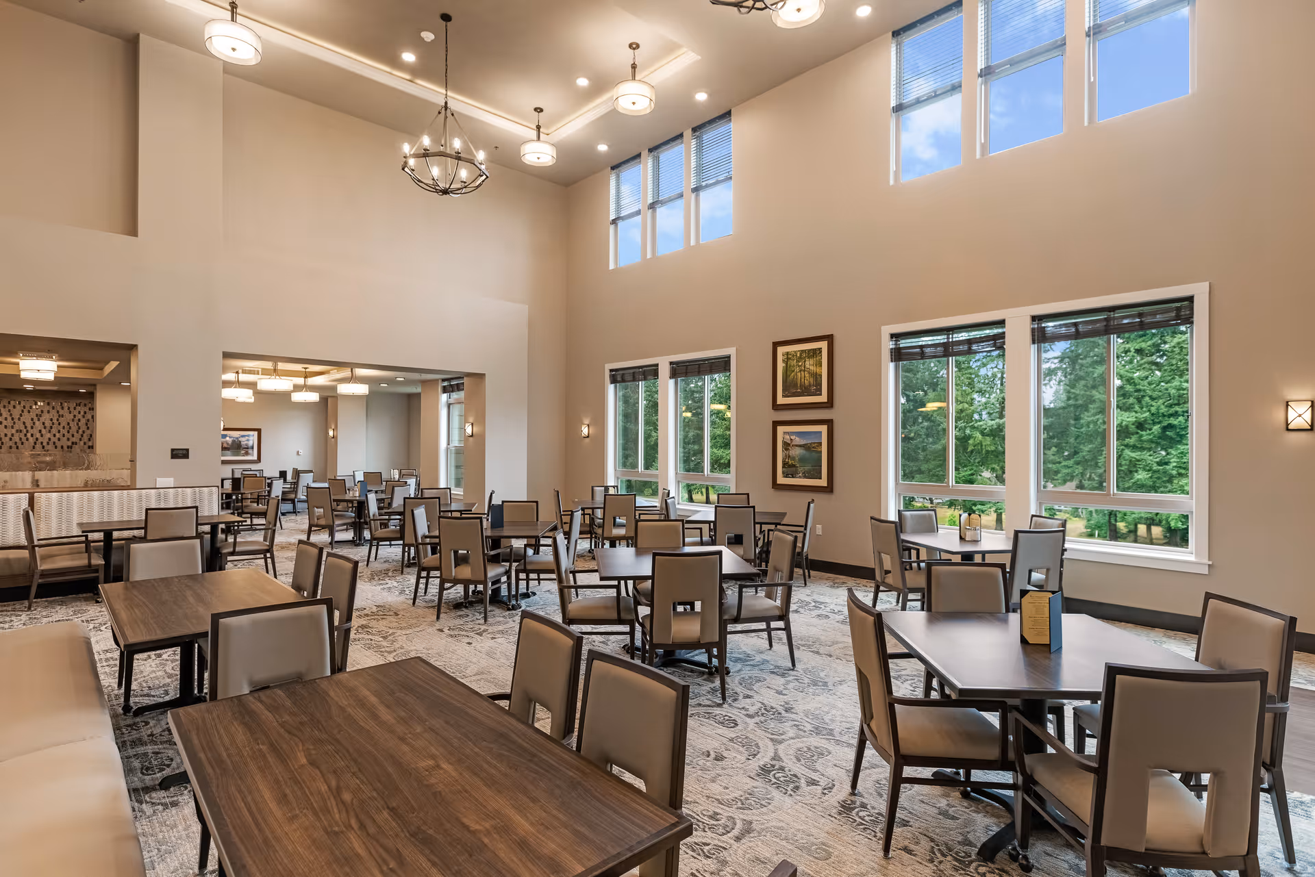 Spacious, well-lit dining room with multiple tables and chairs, high ceilings, chandeliers, and large windows overlooking trees.
