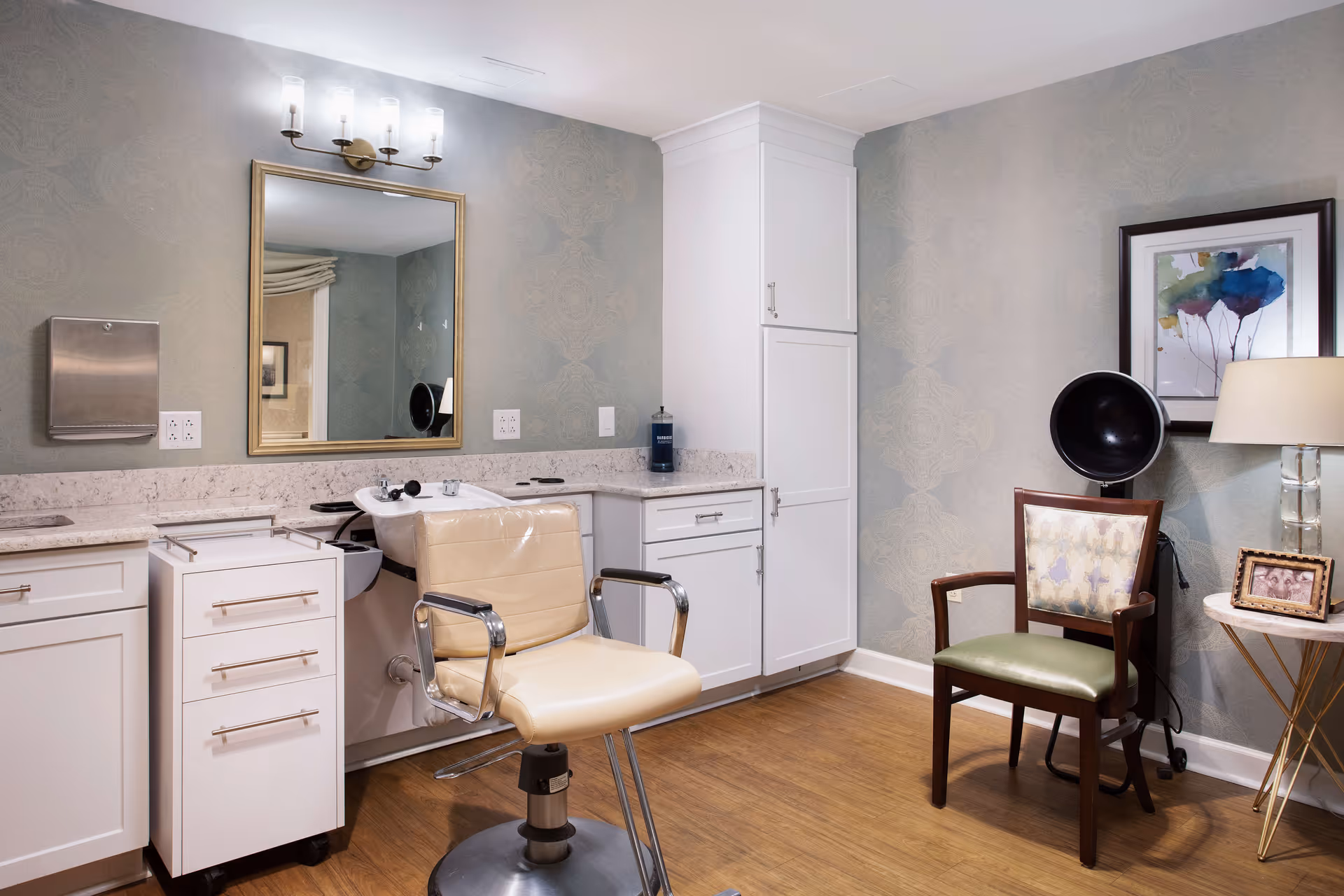 A small salon/hairdressing room with a styling chair and wash sink in front of a mirror, white cabinets, and a waiting chair with framed art.