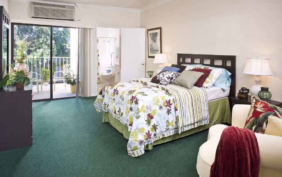 A bright bedroom with a floral patterned bedspread on a bed with multiple pillows. There is a green carpet, a white armchair with a red throw blanket, and a dark dresser with plants on top. A sliding glass door leads to a balcony with outdoor chairs and plants. An open door reveals a bathroom with a toilet and shower.