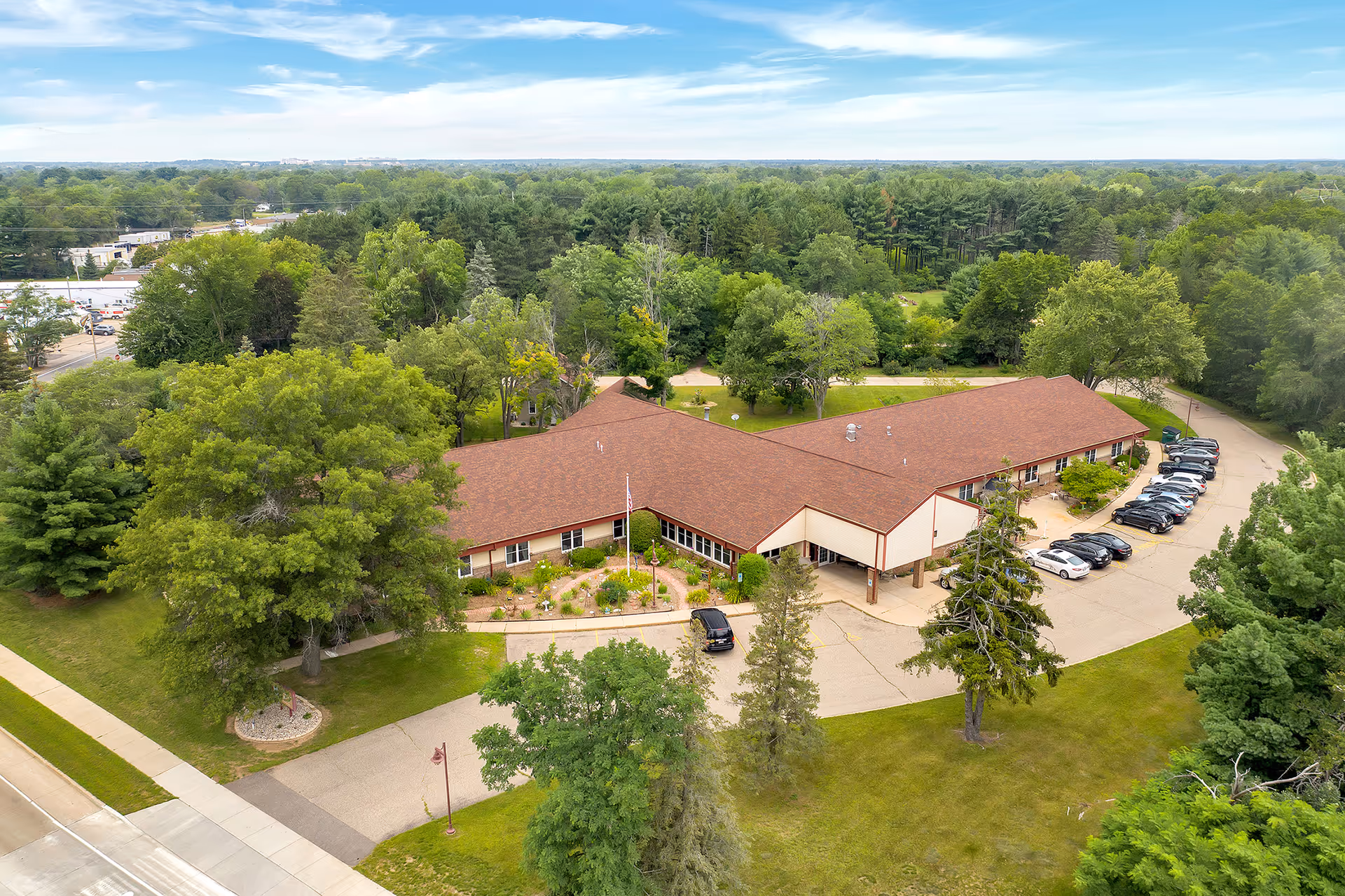 Aerial view of Wellington Place at Whiting, a single-story senior living facility with a brown roof surrounded by lush green trees and landscaped gardens. There is a parking lot with several cars on the right side and a driveway leading to the entrance. The building is set in a spacious, green area with a clear blue sky above.