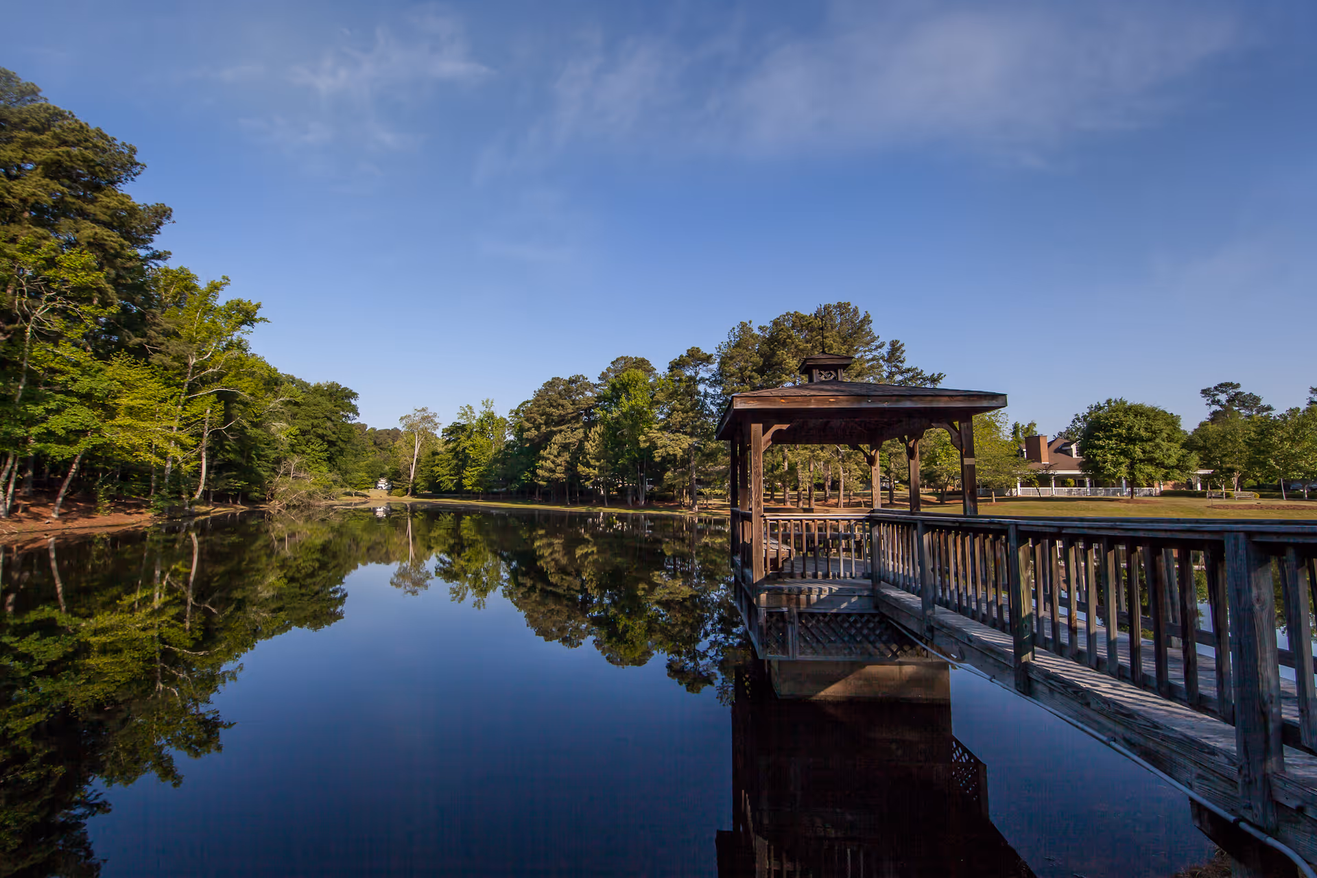 Wooden gazebo and dock extending over a calm reflective lake surrounded by trees under a blue sky.