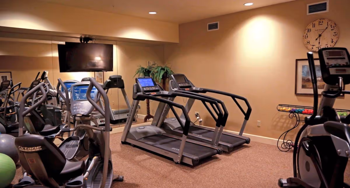 Indoor fitness room with exercise equipment including two treadmills, an elliptical machine, and a stationary bike. A large mirror covers one wall, reflecting the equipment. A flat-screen TV is mounted on the wall above the treadmills. There is a clock and framed pictures on the wall, and a rack holding colorful small weights.