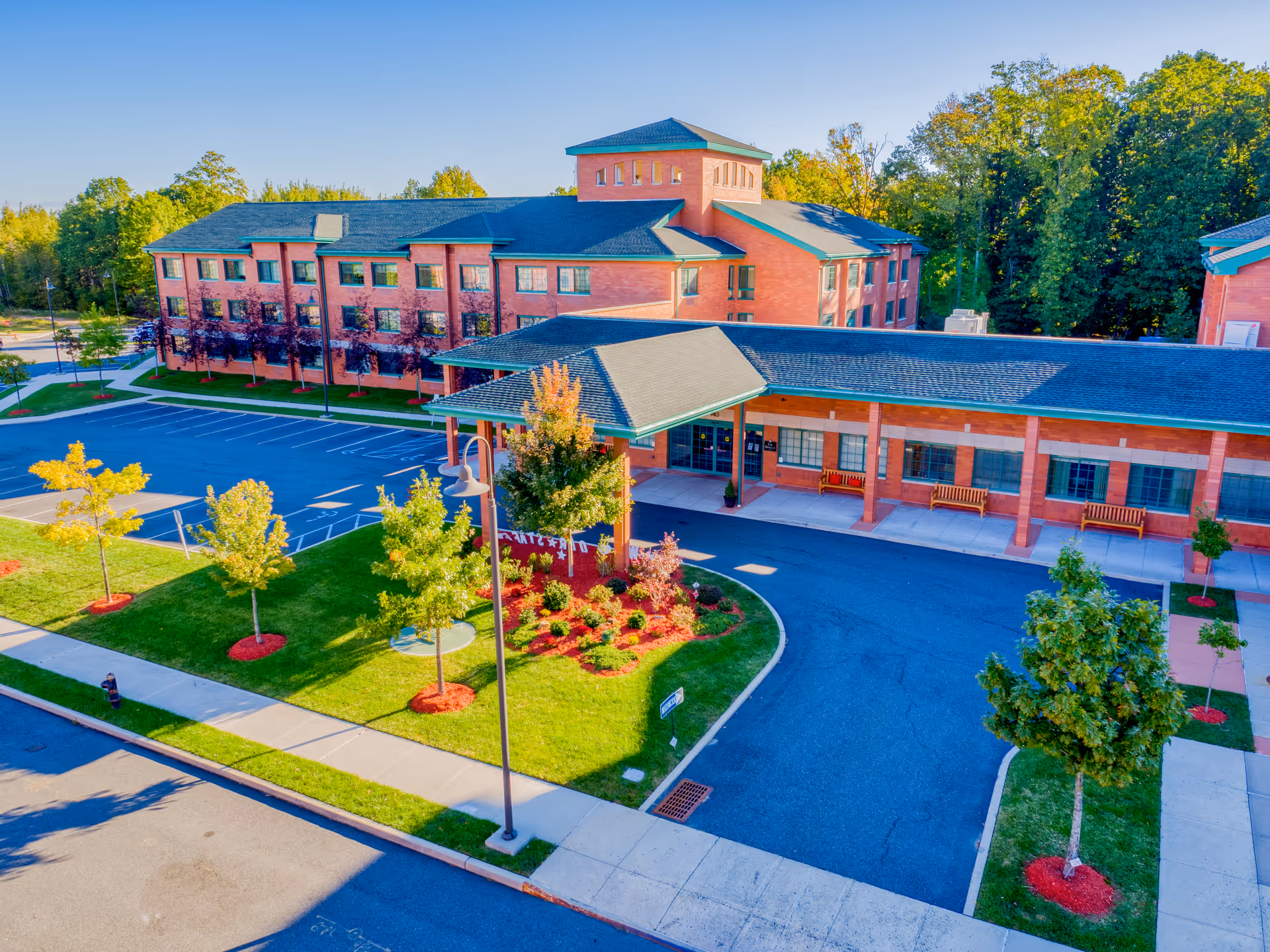 Aerial view of a brick senior living building with a covered entrance, landscaped grounds, and a nearby parking lot.