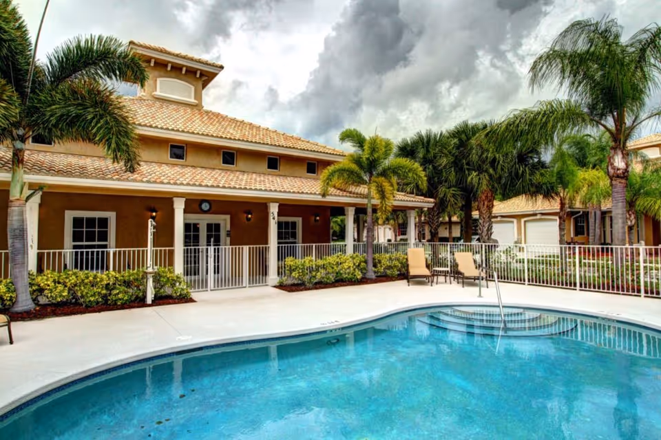 Outdoor swimming pool and lounge area with palm trees in front of a Mediterranean-style clubhouse.