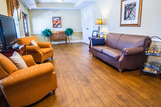 A cozy living room area with wooden flooring, featuring a brown leather sofa on the right, two orange upholstered armchairs on the left, a small wooden side table with a lamp, framed artwork on the walls, a TV on a stand, and two green potted plants near a wooden bench against the far wall.