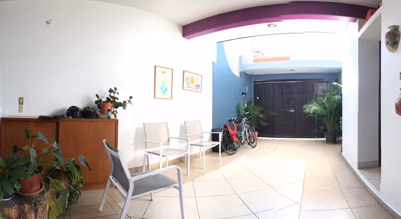 A covered outdoor patio area with white walls and tiled floor. There are several chairs arranged along the wall, a wooden cabinet with potted plants and helmets on top, and two bicycles parked near a black gate. The area is decorated with framed pictures on the wall and some potted plants.