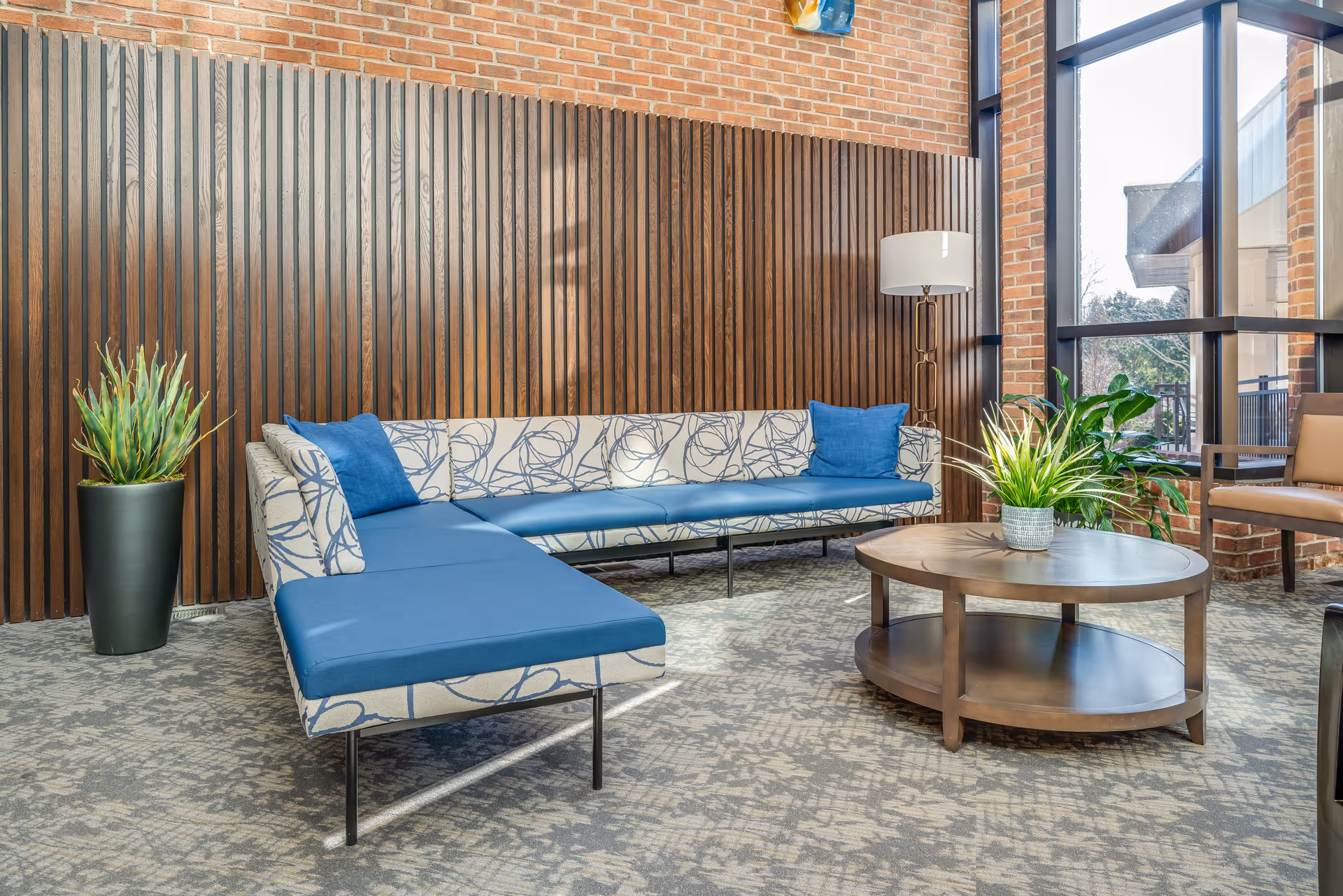 A modern lounge area with a patterned sectional sofa featuring blue cushions, a round wooden coffee table with a potted plant, a tall floor lamp, and large windows letting in natural light. The background includes a brick wall and vertical wooden slats, with additional plants placed around the seating area.