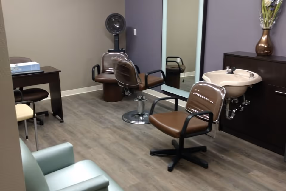 Small salon room with brown styling chairs, a wash basin, large mirror, hooded dryer, and seating.