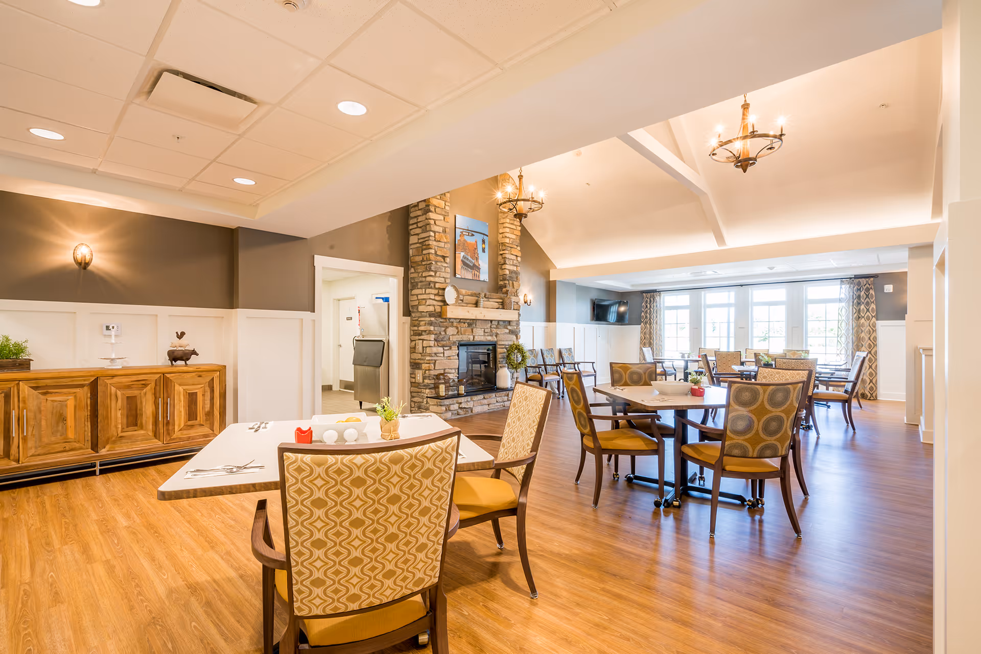 A bright and spacious dining room in a senior living facility with wooden floors, multiple tables and chairs with patterned upholstery, a stone fireplace, and large windows letting in natural light. The room features warm lighting fixtures and a wooden sideboard against one wall.