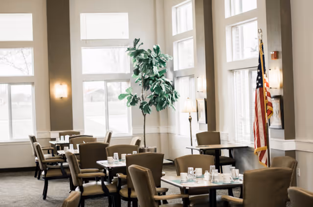 Bright dining room with multiple set tables and chairs, tall windows, a potted tree, and an American flag.