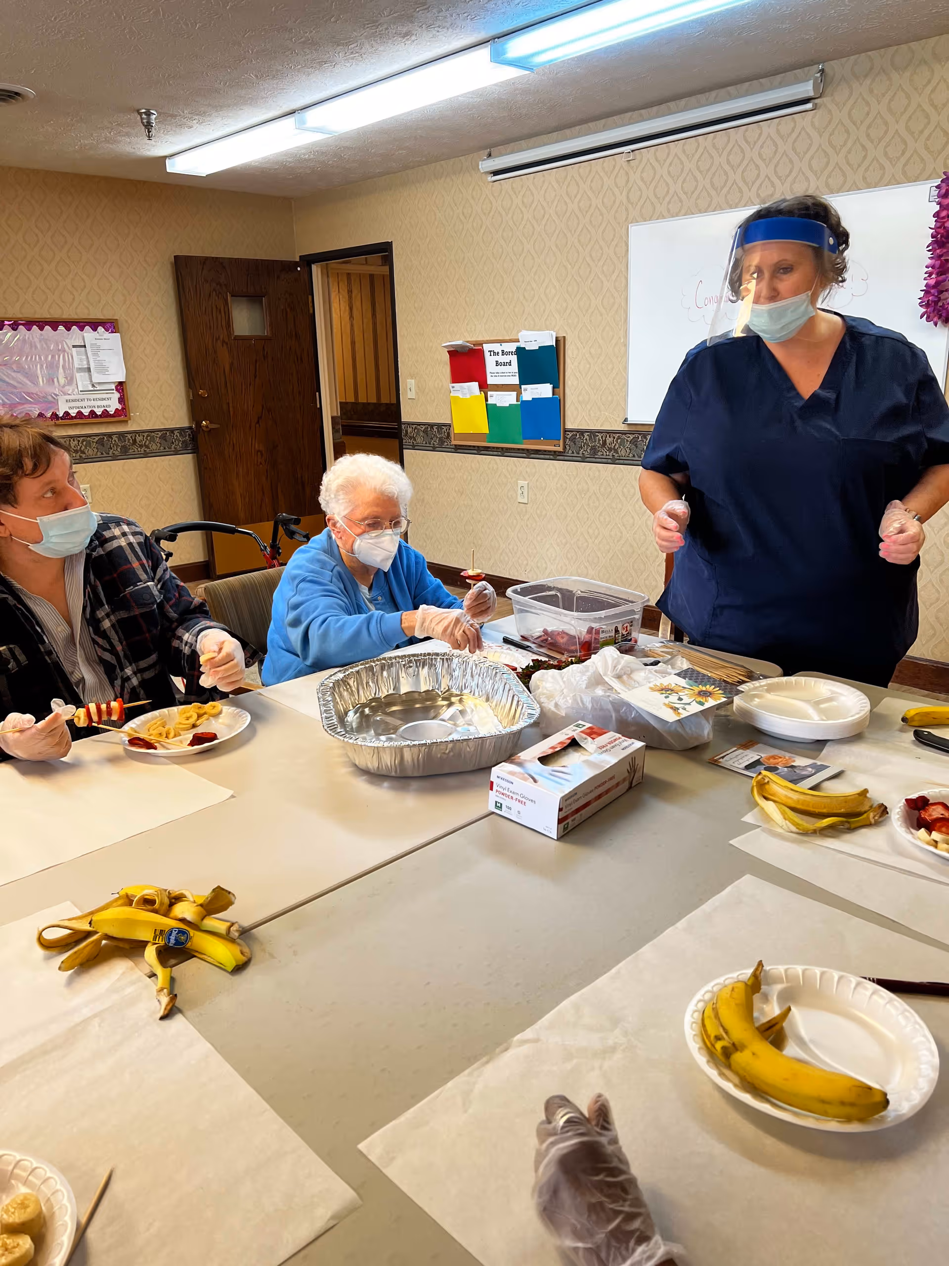 Three people wearing masks and gloves are seated around a table in a room with beige wallpaper. Two elderly individuals are preparing fruit skewers with bananas and strawberries, while a staff member in navy scrubs and a face shield stands nearby. The table has paper plates with bananas, a box of gloves, and a large aluminum tray.
