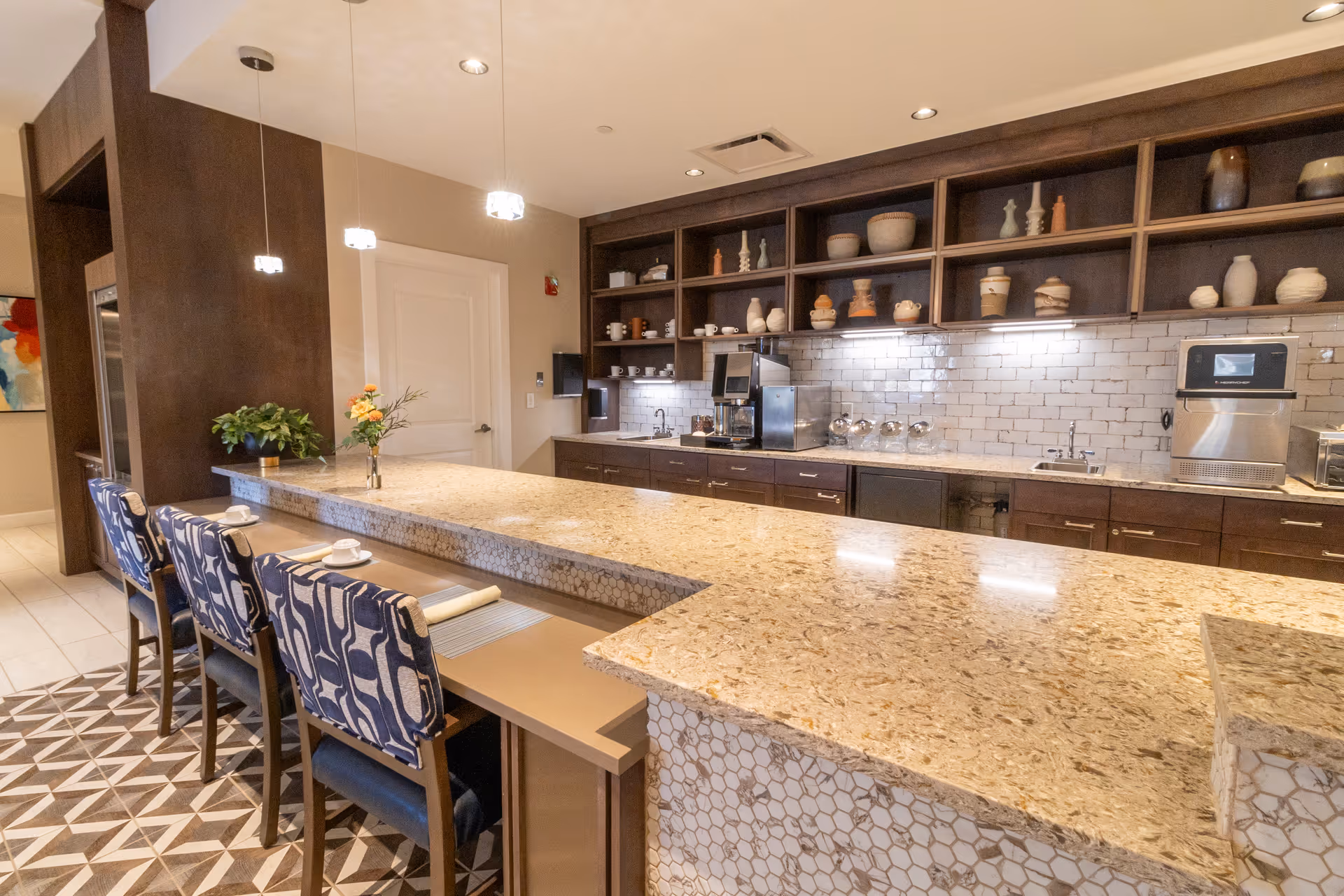 Long granite-topped communal kitchen/bar with patterned bar stools, open shelving, and stainless appliances.