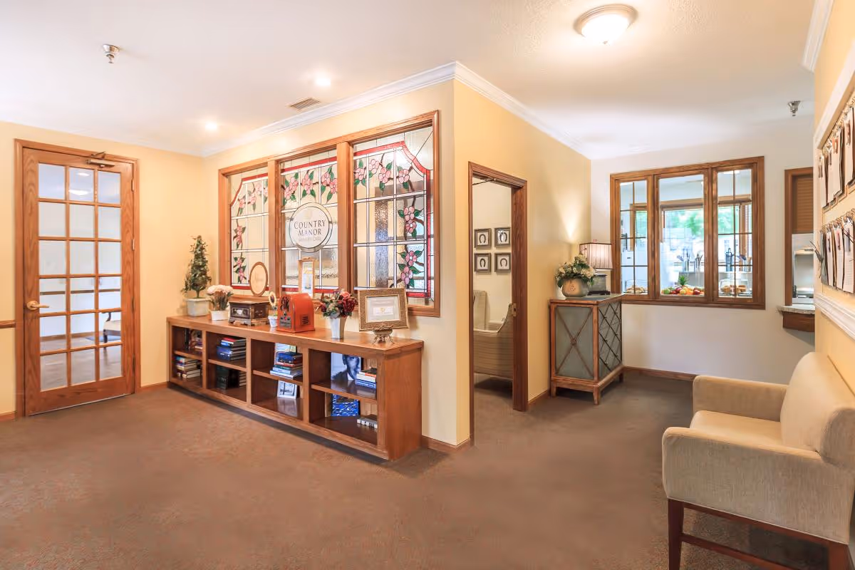 A cozy interior space with beige walls and carpeted floor featuring a wooden door with glass panels on the left, a wooden shelf with books and decorative items beneath stained glass windows that say 'Country Manor', a small cabinet with a lamp and flowers, and a beige armchair on the right.
