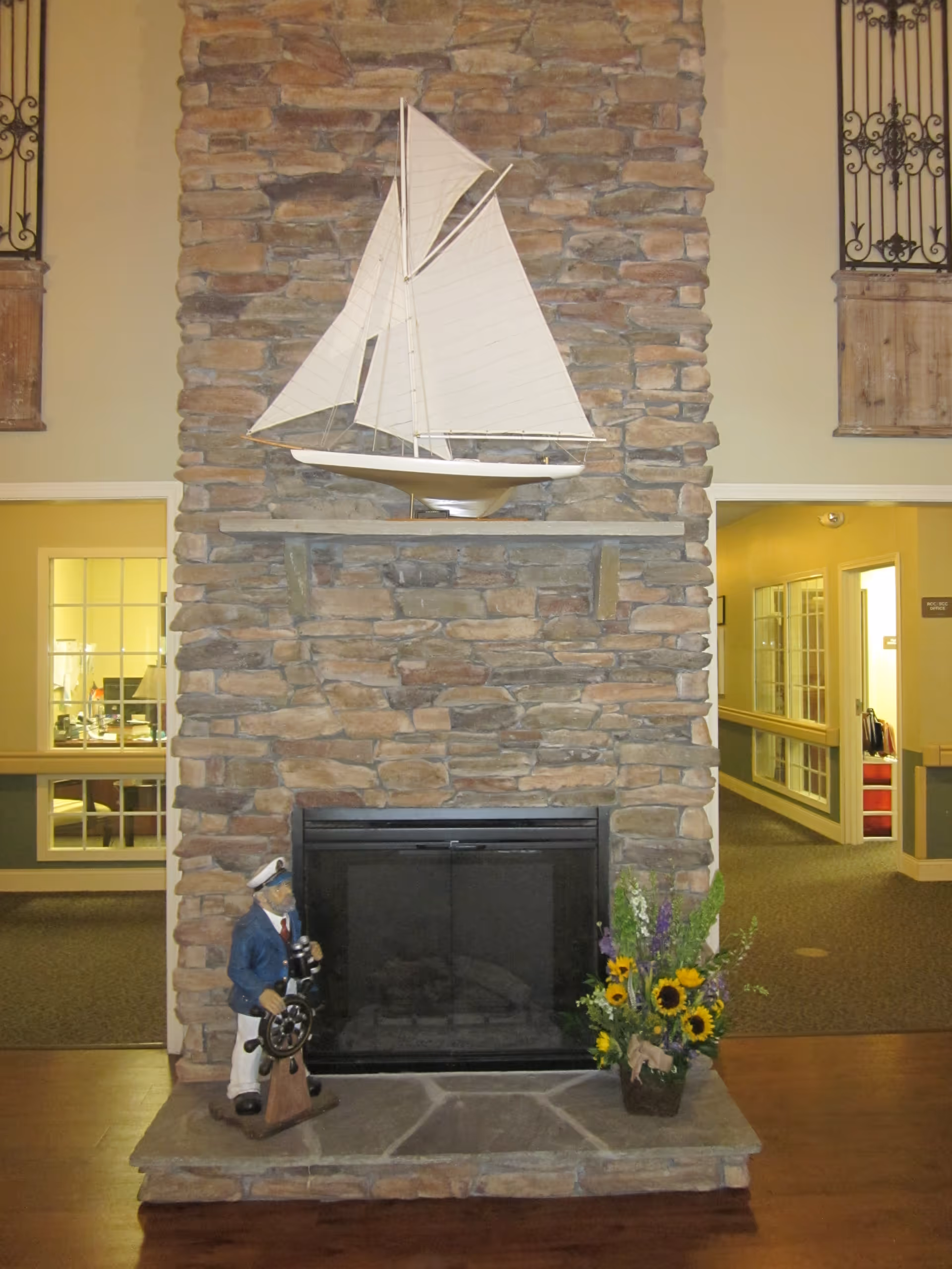 Stone fireplace with a model sailboat on the mantle, a nautical figurine and a sunflower arrangement on the hearth, flanked by interior hallways.