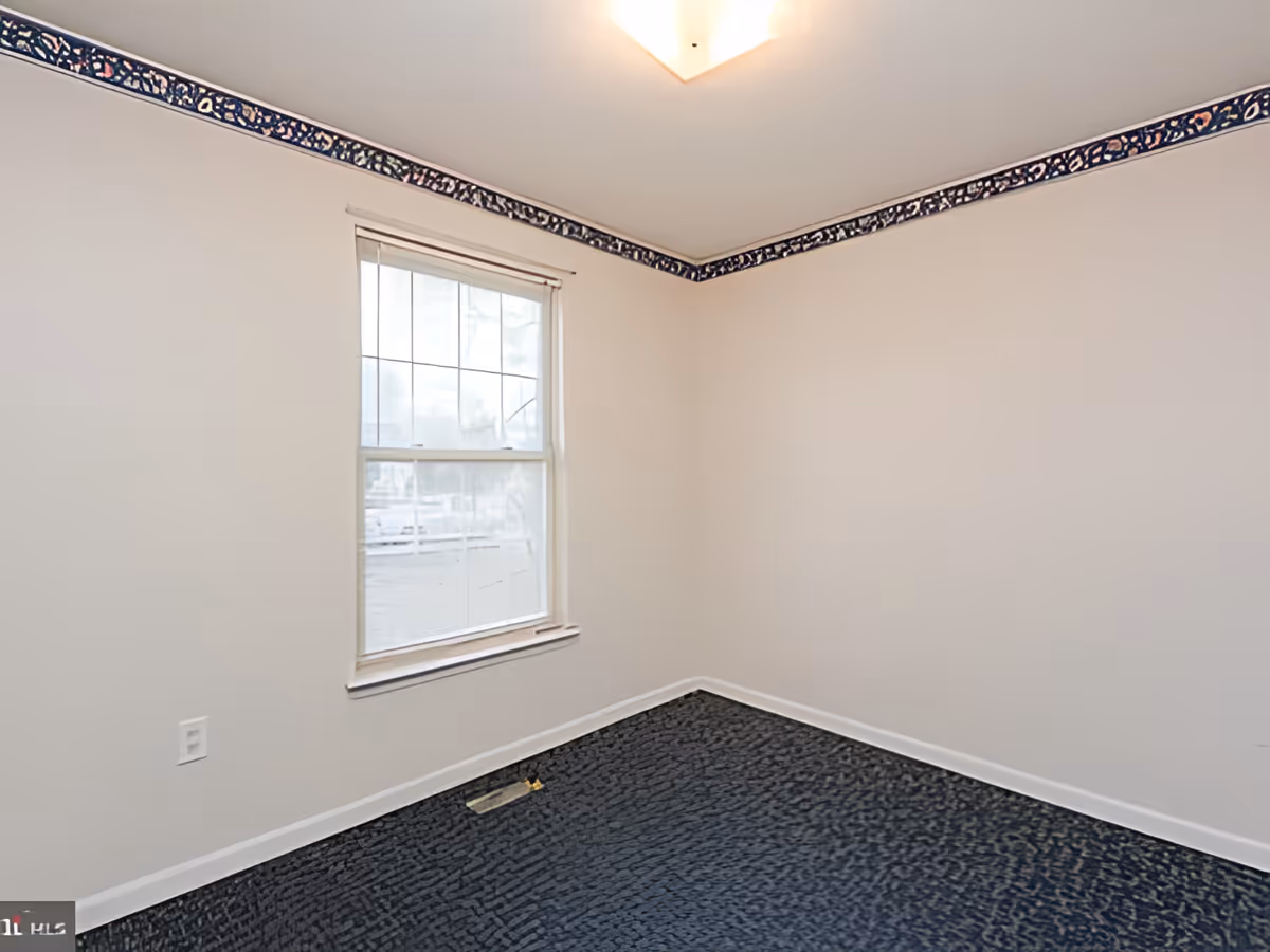 Empty room with beige walls, a single window with blinds, a dark patterned carpet, and a decorative border near the ceiling.