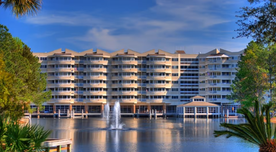 A large multi-story residential building with balconies overlooking a pond with a water fountain in the center. The building is surrounded by trees and greenery under a partly cloudy blue sky.