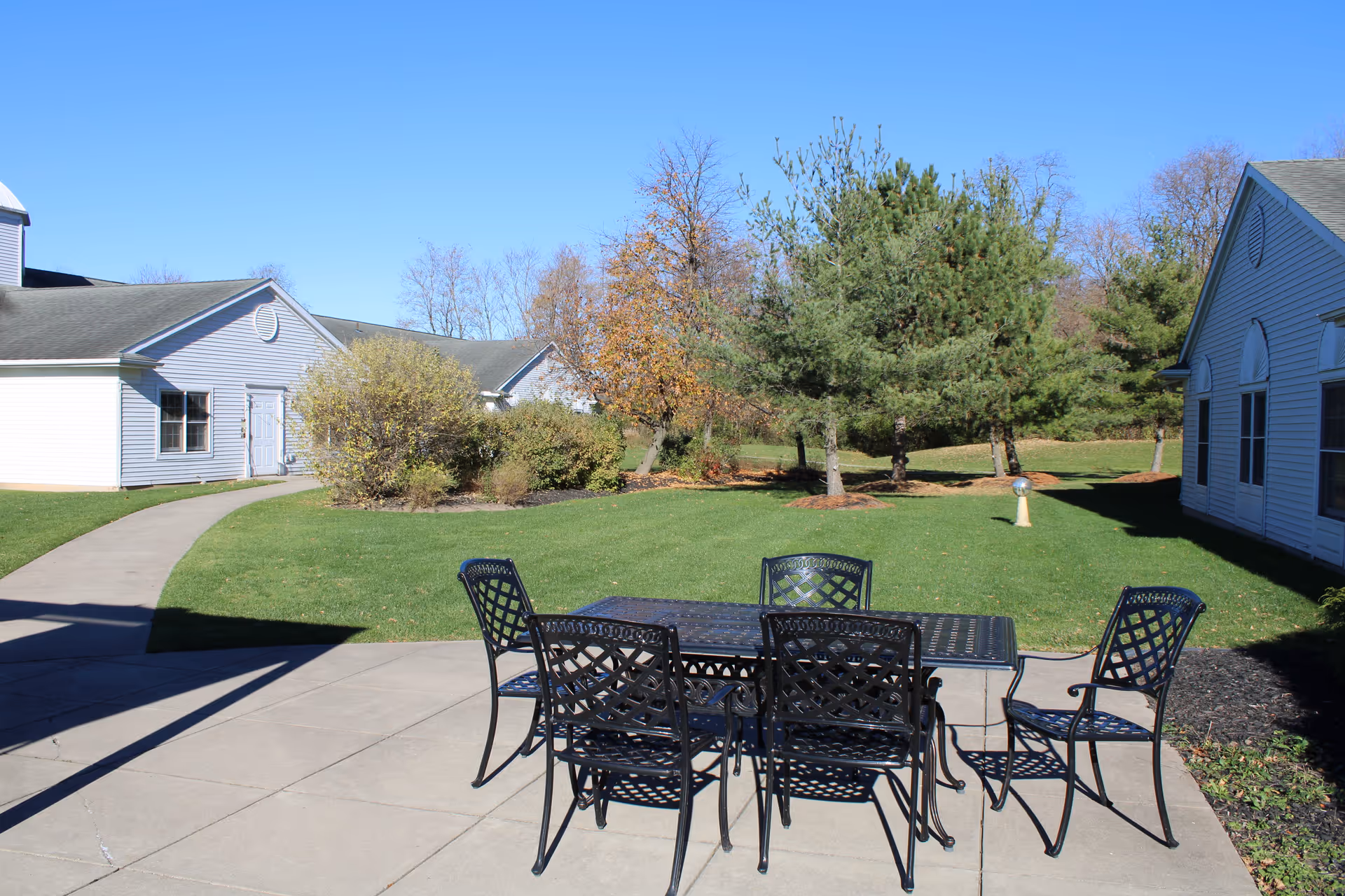 Outdoor patio area with a black metal table and six matching chairs on a concrete surface. Surrounding the patio is a well-maintained green lawn with trees and shrubs. Two light-colored buildings with gray roofs are visible on either side of the patio under a clear blue sky.