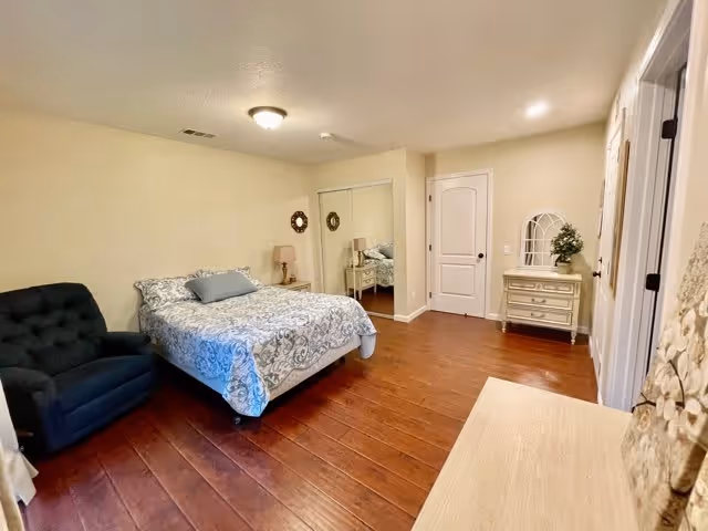 A bedroom with a bed covered in a patterned blue and white comforter, a dark blue armchair to the left, wooden flooring, a dresser with a small plant and decorative mirror on the right, and a closet with mirrored sliding doors.