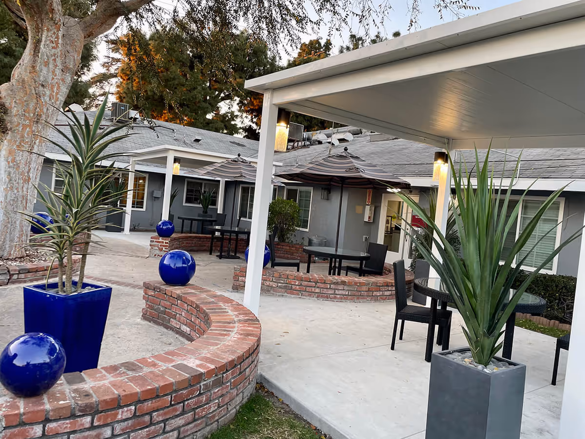 Outdoor patio area at Whittier Pacific Center featuring brick curved seating walls with blue decorative spheres, potted plants, tables with black chairs, and striped umbrellas. The building exterior is visible in the background with windows and doors under a covered walkway.