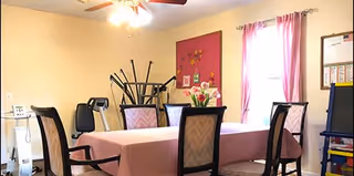 A room with a rectangular table covered with a pink tablecloth surrounded by six chairs. The room has beige walls, a ceiling fan with lights, a window with pink curtains, a bulletin board, and some stacked chairs in the corner. There are also flowers on the table and a few items on a small table near the wall.