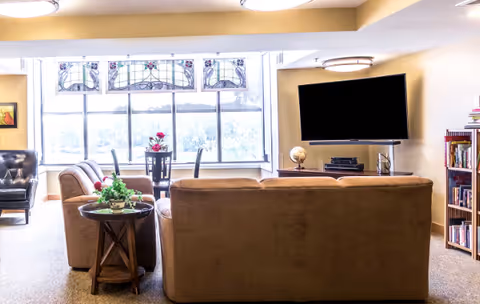 Sunlit communal living room with sofas facing a wall-mounted TV and a windowed dining area.