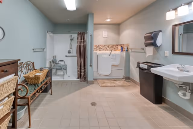 A spacious, accessible bathroom featuring a walk-in shower with a shower chair and grab bars, a walk-in bathtub with a safety handle, a wall-mounted sink with a mirror above, a paper towel dispenser, a trash bin, and a bench with baskets for storage. The walls are painted light blue and the floor is tiled.