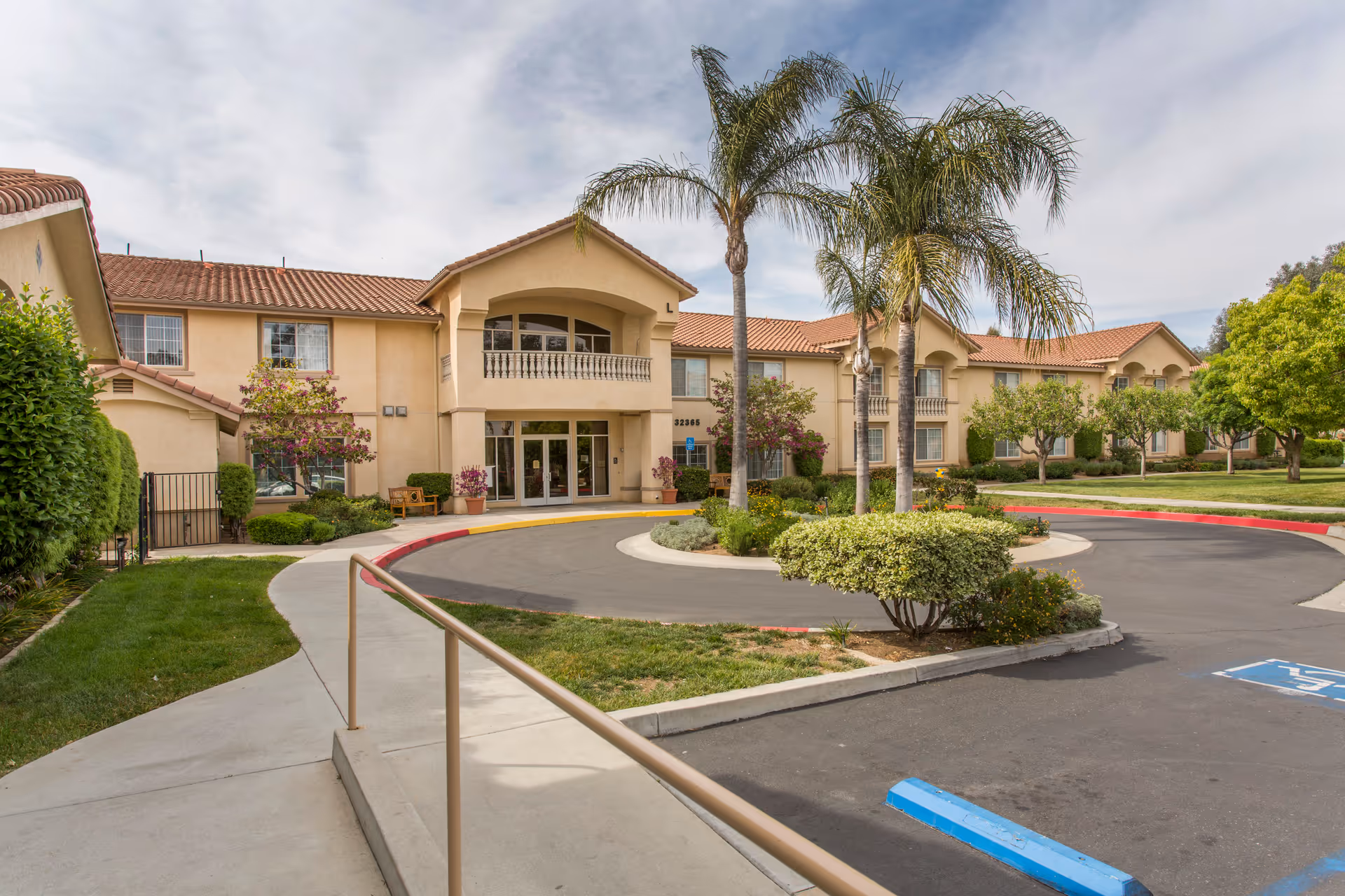 Exterior view of Wildomar Senior facility showing a two-story beige building with a red tile roof, palm trees, bushes, and a circular driveway with a sidewalk and handicap parking space in front.